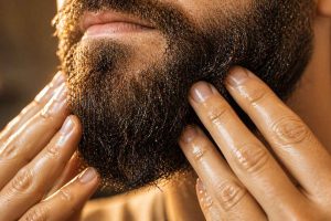 Gros plan des mains d'un homme massant de l'huile à barbe dans une barbe épaisse, depuis la peau jusqu'aux pointes. Technique d'application correcte pour une barbe nourrie